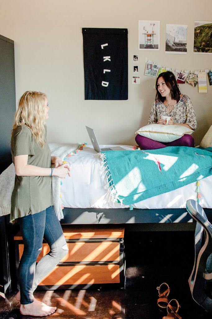Two female students are in a dorm room talking. One is standing up, and the other is sitting on her bed.